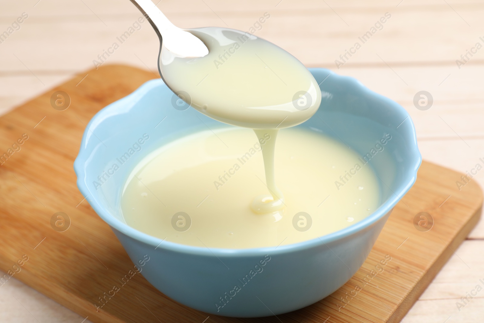 Photo of Condensed milk flowing down from spoon into bowl on wooden table, closeup