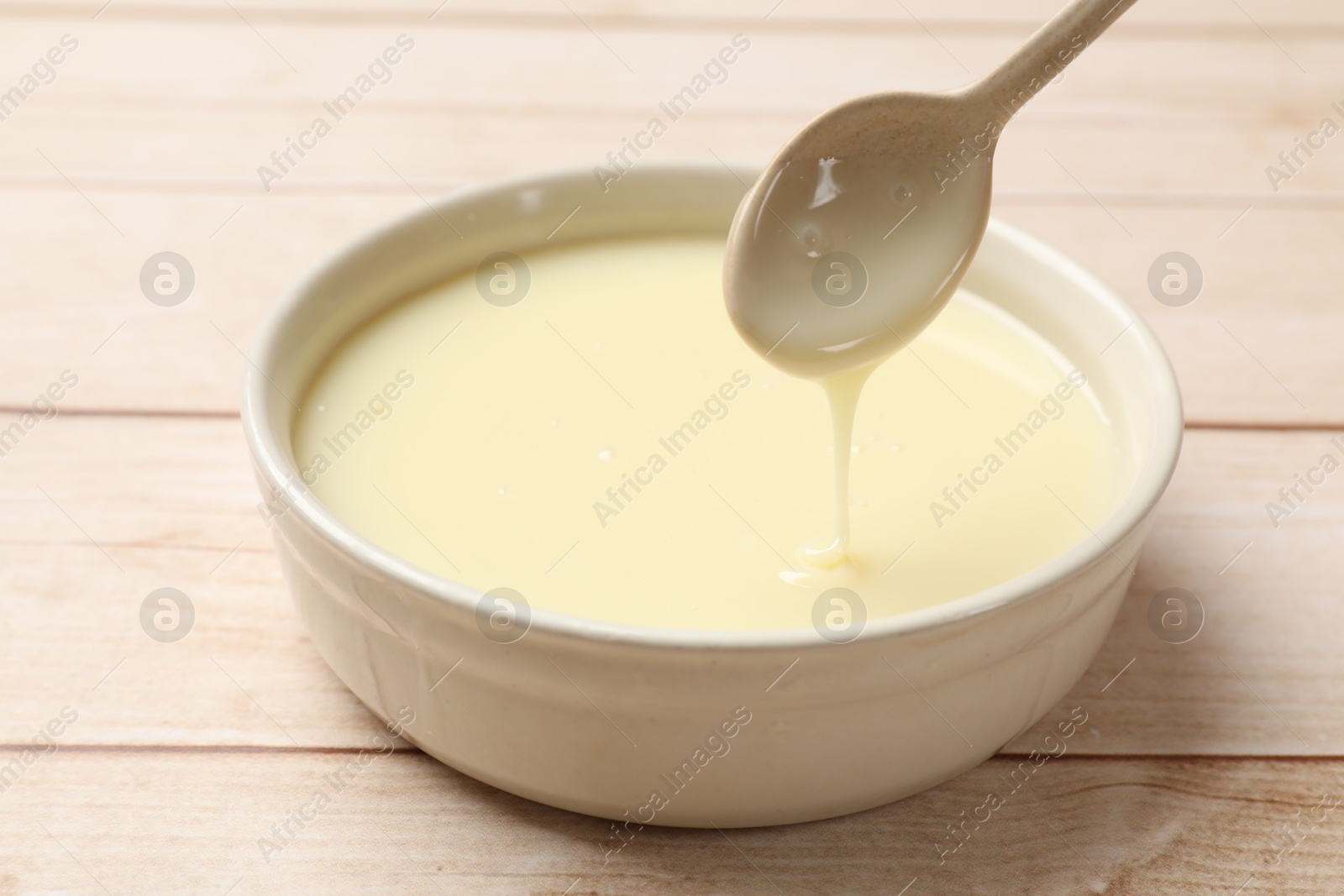 Photo of Condensed milk flowing down from spoon into bowl on wooden table, closeup