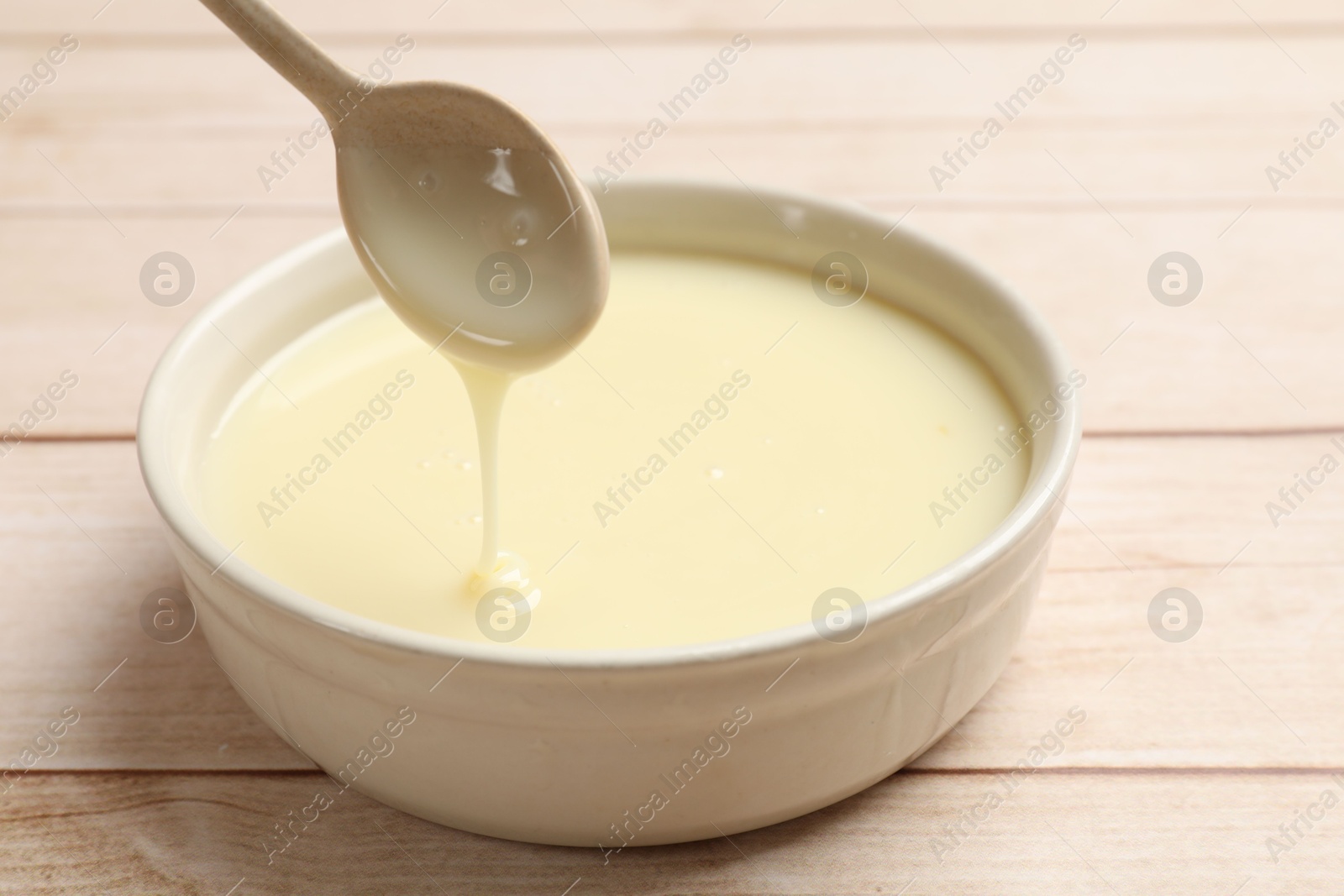 Photo of Condensed milk flowing down from spoon into bowl on wooden table, closeup