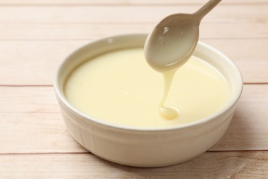 Photo of Condensed milk flowing down from spoon into bowl on wooden table, closeup