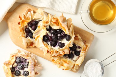 Photo of Delicious puff pastries with blueberries, powdered sugar, honey and tea on white table, flat lay