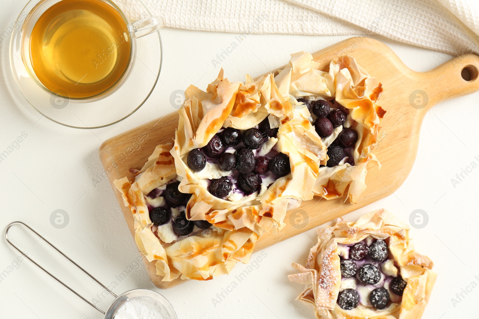 Photo of Delicious puff pastries with blueberries, powdered sugar, honey and tea on white table, flat lay