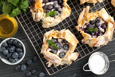 Photo of Tasty puff pastries with blueberries, powdered sugar, mint and honey on dark table, flat lay