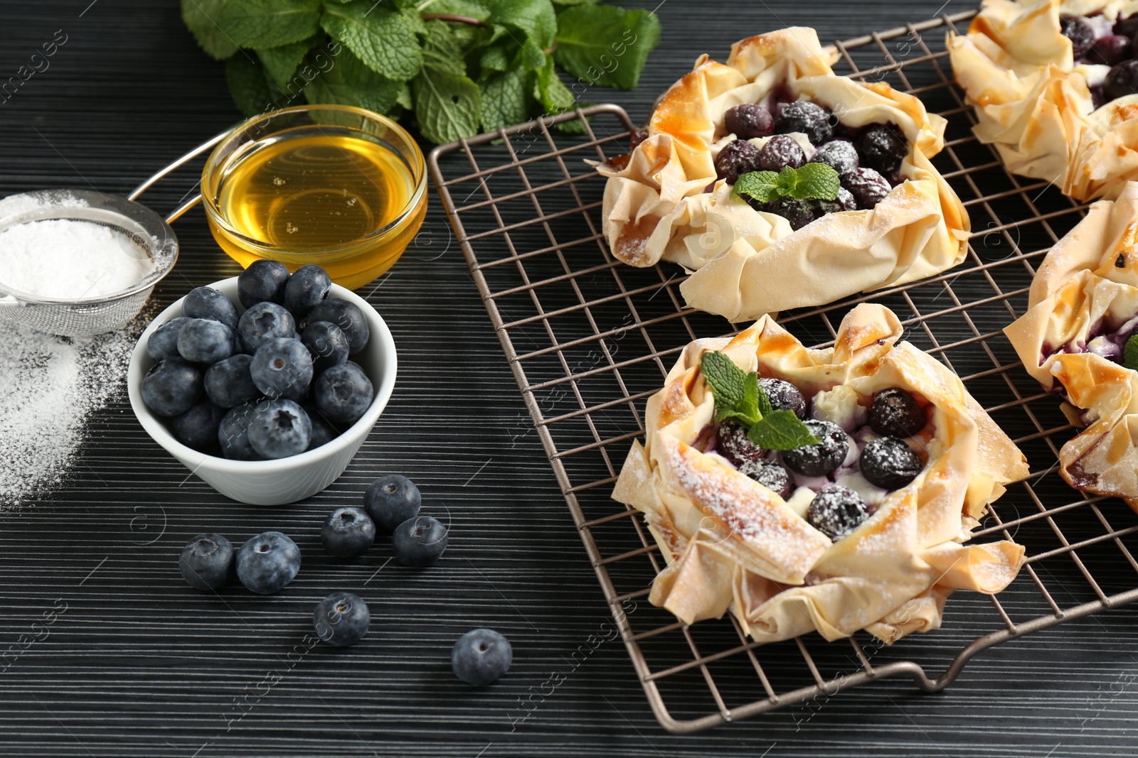 Photo of Tasty puff pastries with blueberries, powdered sugar, mint and honey on dark table, closeup