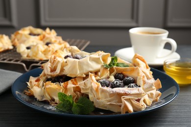 Photo of Tasty puff pastries with blueberries, powdered sugar, mint, coffee and honey on dark table, closeup