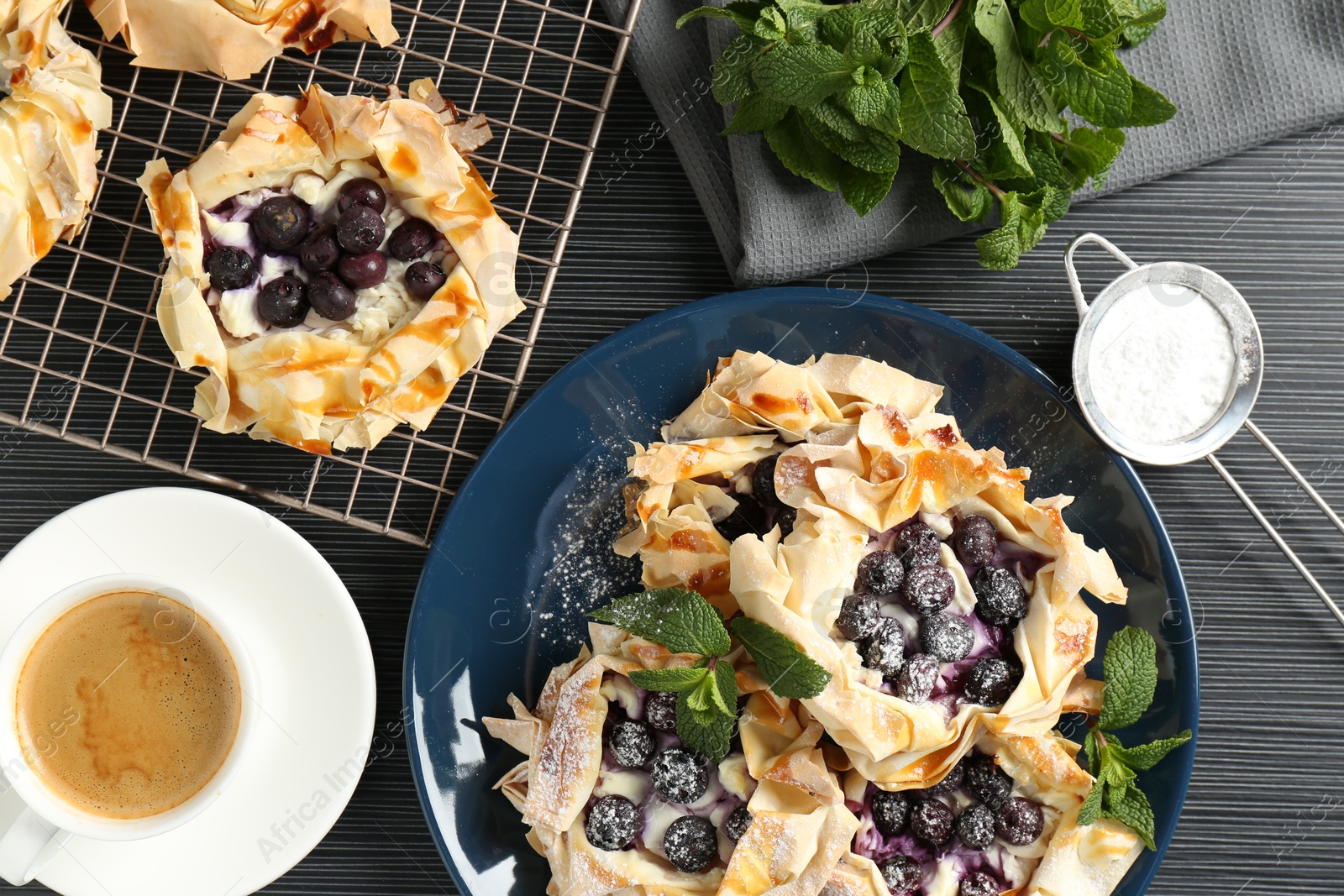 Photo of Tasty puff pastries with blueberries, powdered sugar, mint and coffee on dark table, flat lay