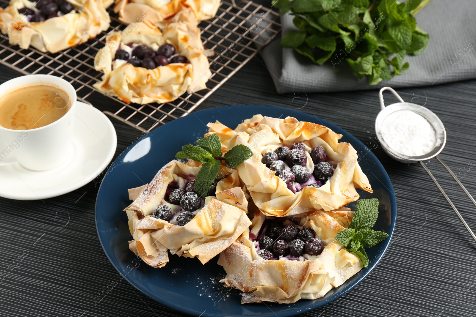 Photo of Tasty puff pastries with blueberries, powdered sugar, mint and coffee on dark table, closeup