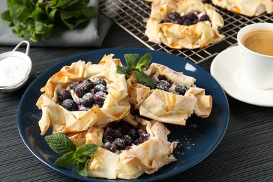 Photo of Tasty puff pastries with blueberries, powdered sugar, mint and coffee on dark table, closeup