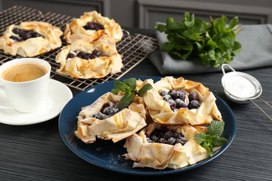 Photo of Tasty puff pastries with blueberries, powdered sugar, mint and coffee on dark table, closeup