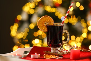 Photo of Aromatic mulled wine in glass cup and burning candles on tray against blurred Christmas lights, bokeh effect