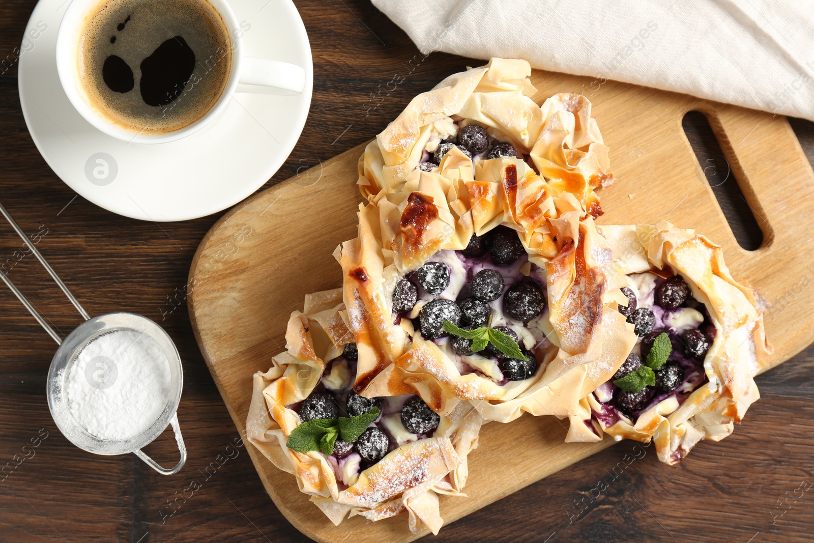 Photo of Tasty puff pastries with blueberries, powdered sugar, mint and coffee on wooden table, flat lay