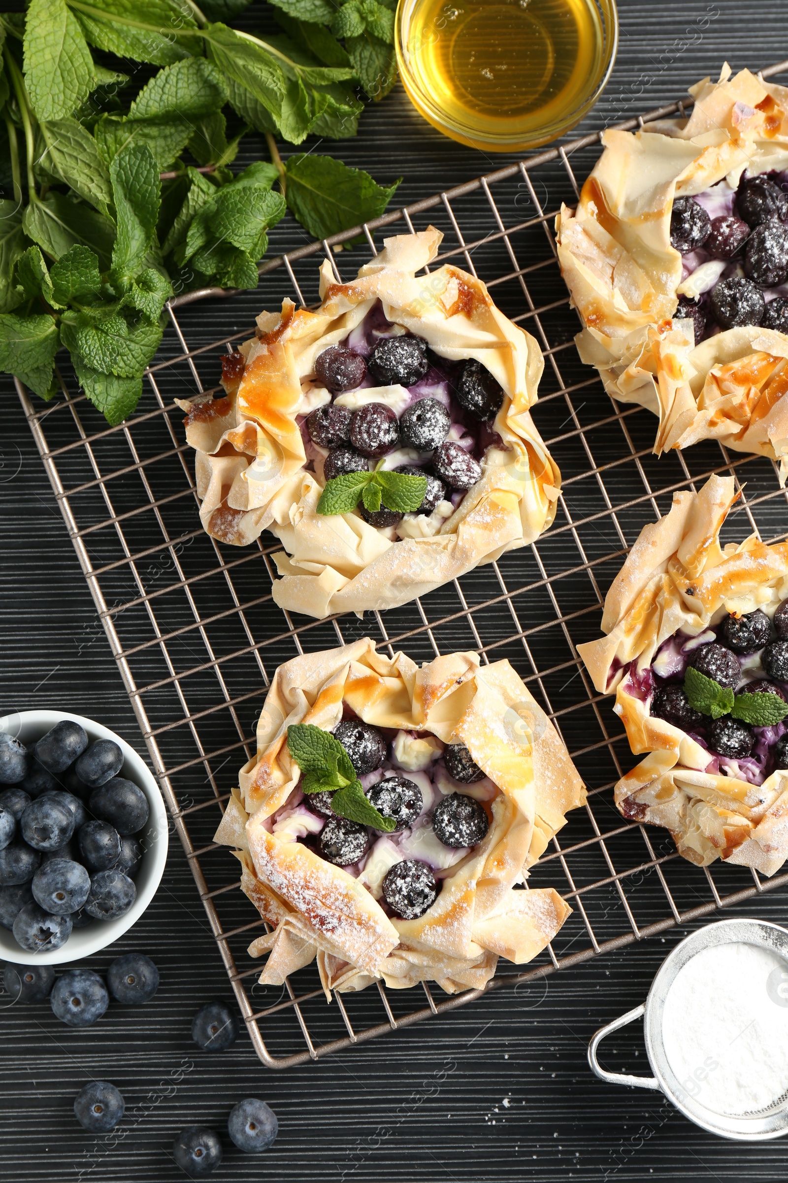 Photo of Tasty puff pastries with blueberries, powdered sugar, mint and honey on dark table, flat lay