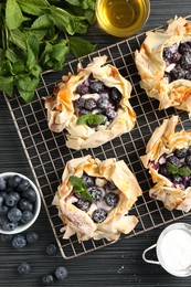 Photo of Tasty puff pastries with blueberries, powdered sugar, mint and honey on dark table, flat lay