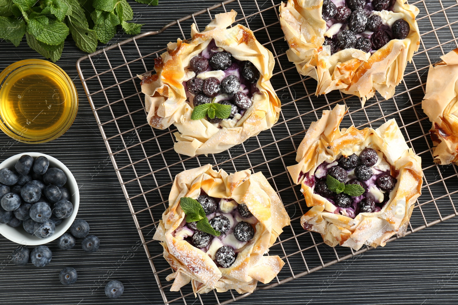 Photo of Tasty puff pastries with blueberries, powdered sugar, mint and honey on dark table, flat lay