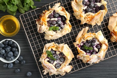 Photo of Tasty puff pastries with blueberries, powdered sugar, mint and honey on dark table, flat lay