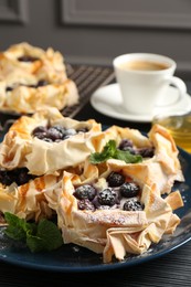 Photo of Tasty puff pastries with blueberries, powdered sugar, mint, coffee and honey on dark table, closeup