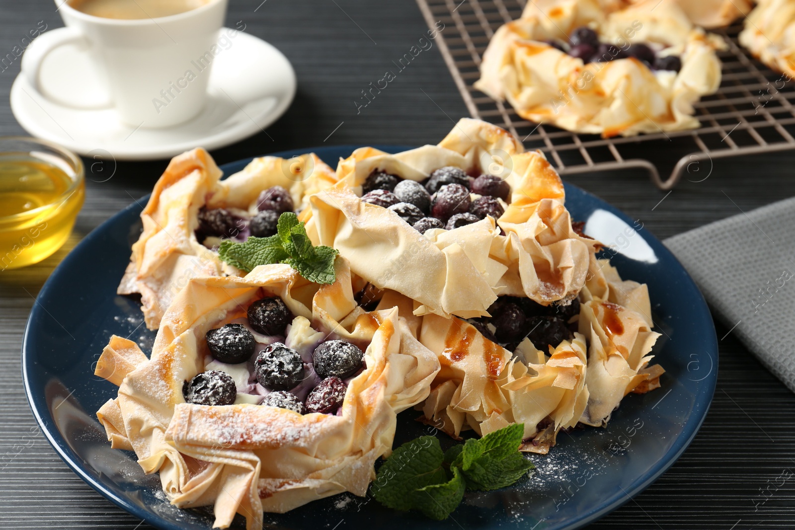 Photo of Tasty puff pastries with blueberries, powdered sugar, mint, coffee and honey on dark table, closeup