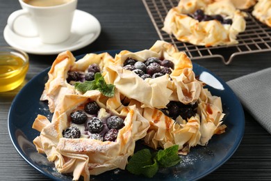 Photo of Tasty puff pastries with blueberries, powdered sugar, mint, coffee and honey on dark table, closeup