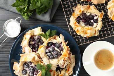 Photo of Tasty puff pastries with blueberries, powdered sugar, mint and coffee on dark table, flat lay