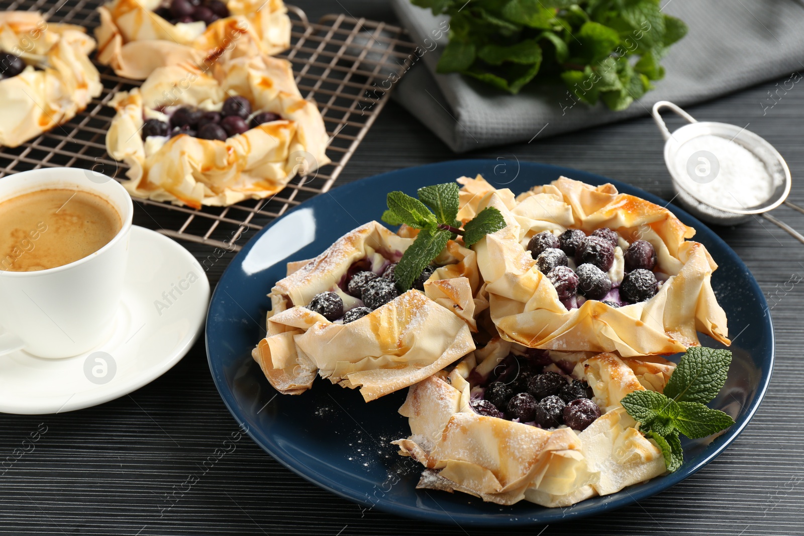 Photo of Tasty puff pastries with blueberries, powdered sugar, mint and coffee on dark table, closeup