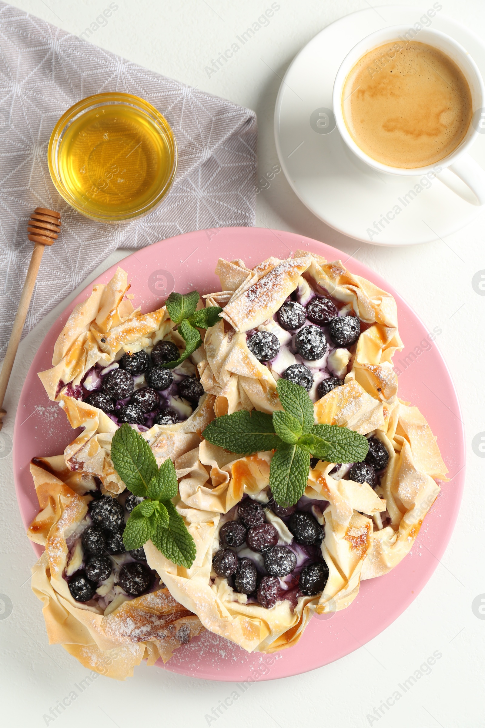 Photo of Tasty puff pastries with blueberries, powdered sugar, mint, honey and coffee on white table, flat lay