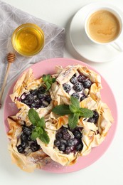 Photo of Tasty puff pastries with blueberries, powdered sugar, mint, honey and coffee on white table, flat lay