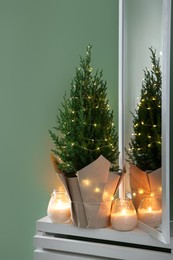 Photo of Small spruce tree decorated with Christmas lights and burning candles on white radiator cabinet near green wall