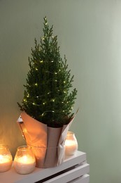 Photo of Small spruce tree decorated with Christmas lights and burning candles on white radiator cabinet near green wall