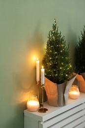 Photo of Small spruce tree decorated with Christmas lights and burning candles on white radiator cabinet near green wall