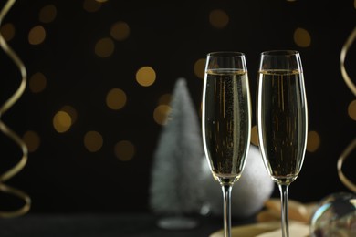 Photo of Delicious champagne in glasses and Christmas decor against black background with blurred lights, closeup. Space for text