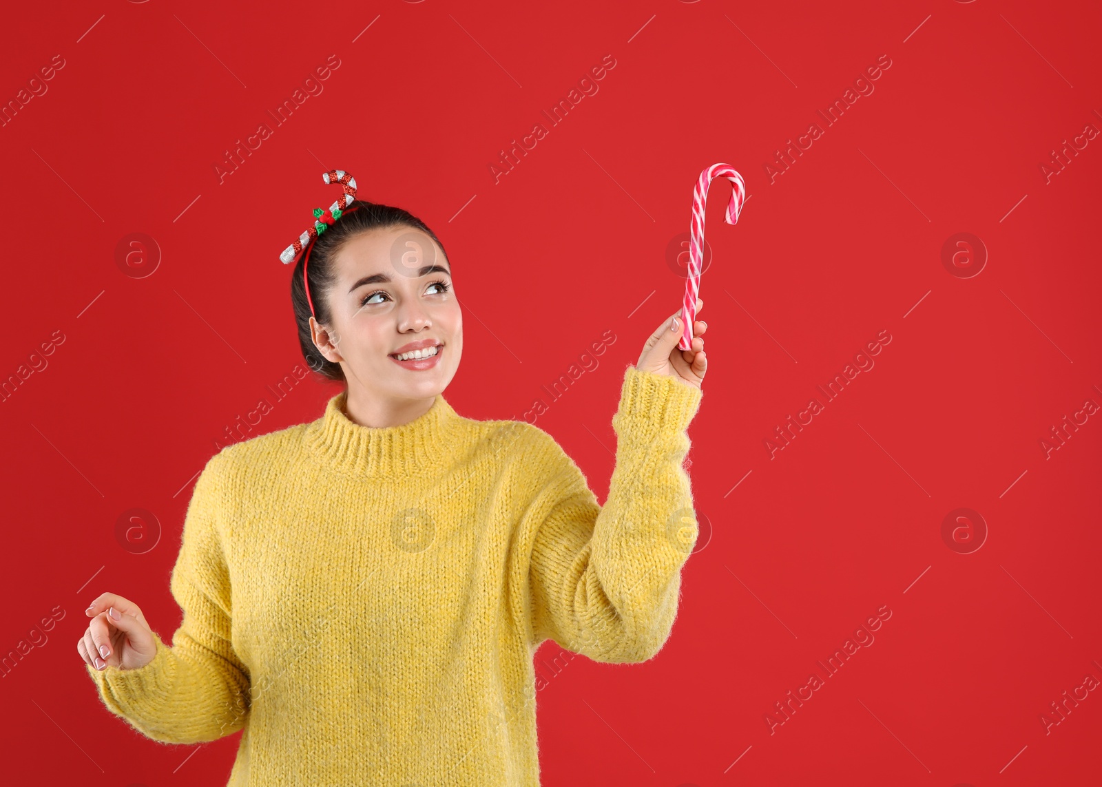 Photo of Young woman in yellow sweater and festive headband holding candy cane on red background, space for text