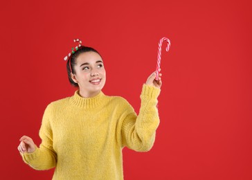 Photo of Young woman in yellow sweater and festive headband holding candy cane on red background, space for text