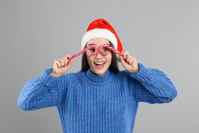 Young woman in blue sweater and Santa hat holding candy canes on grey background. Celebrating Christmas