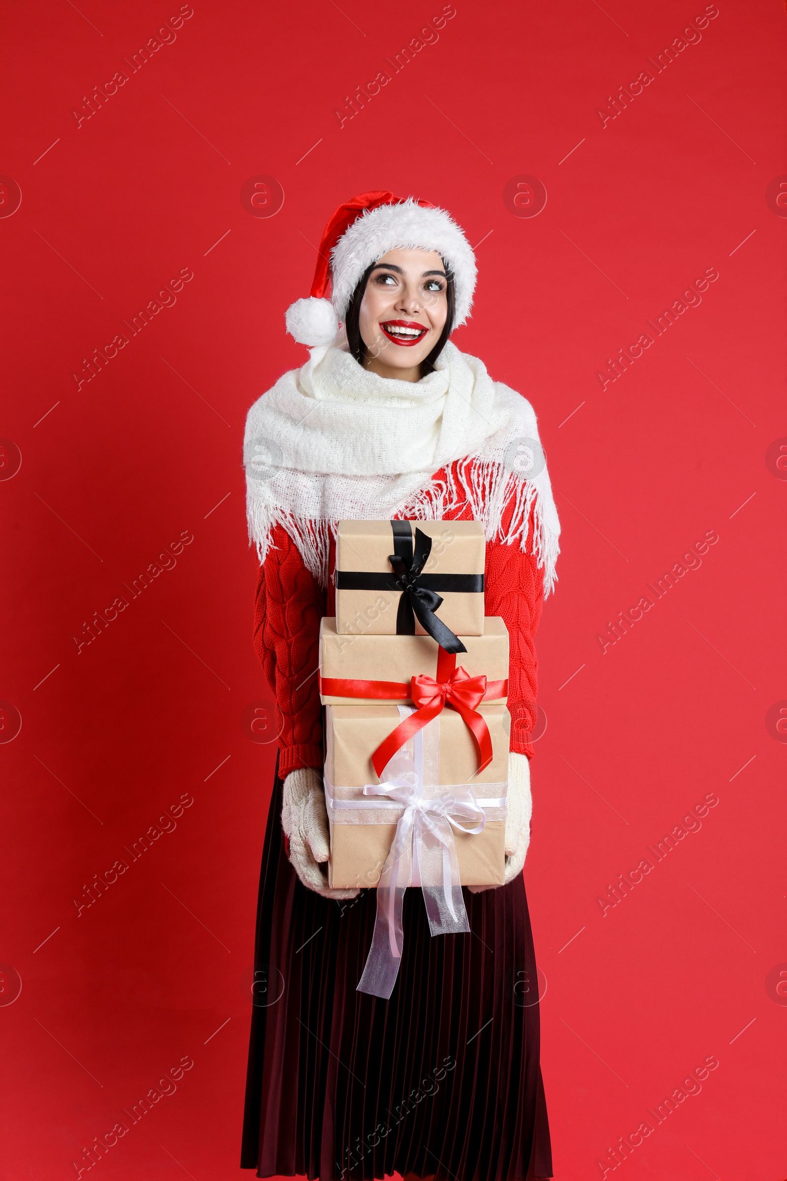 Photo of Woman in Santa hat, scarf and sweater holding Christmas gifts on red background