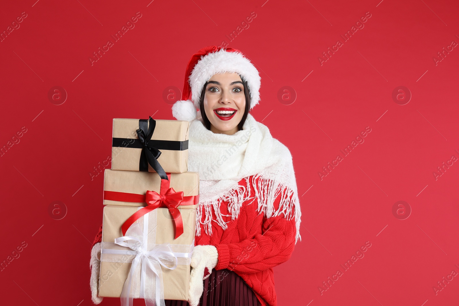 Photo of Woman in Santa hat, scarf and sweater holding Christmas gifts on red background