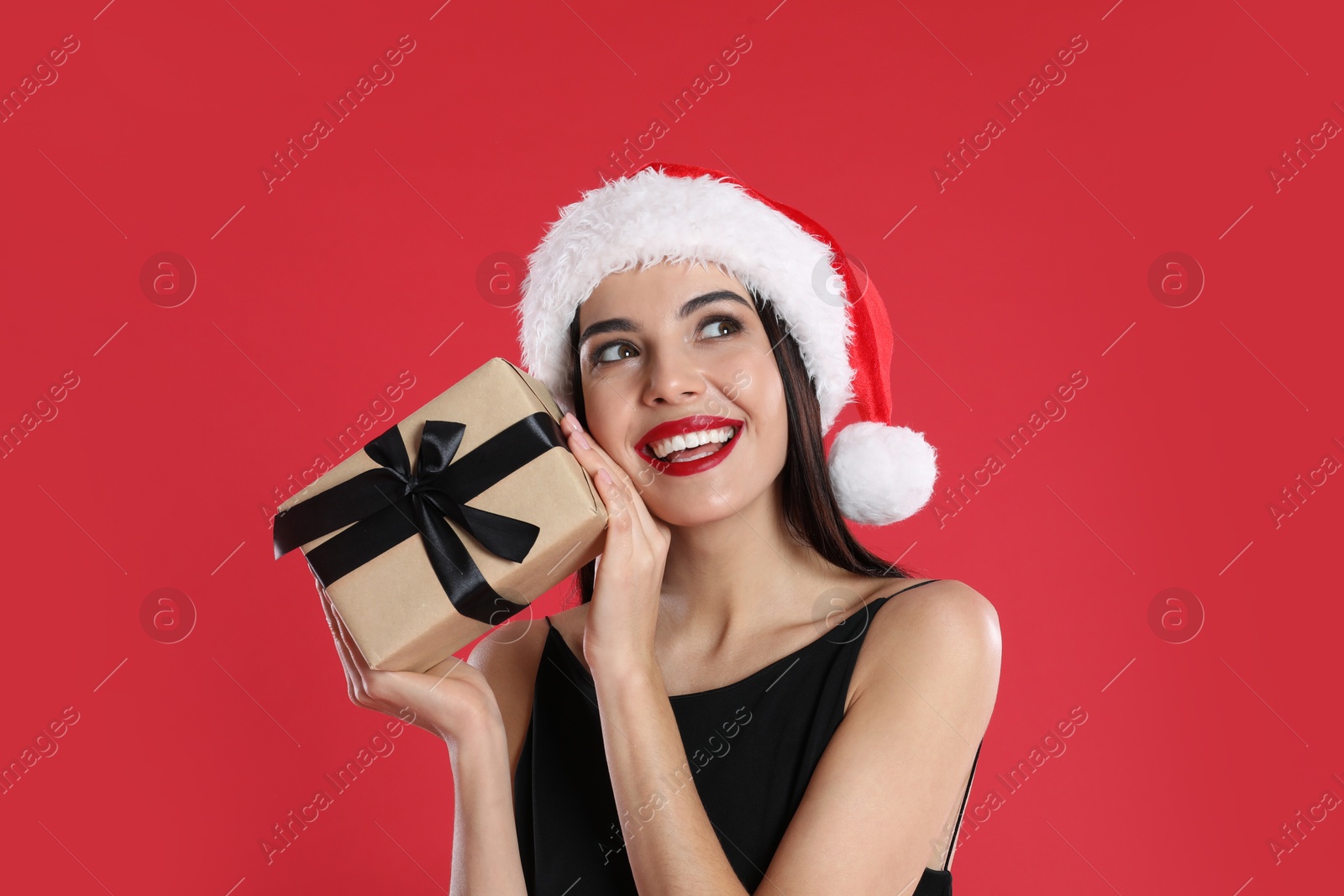 Photo of Woman in black dress and Santa hat holding Christmas gift on red background