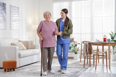 Photo of Caregiver assisting senior woman with walking cane indoors. Home health care service