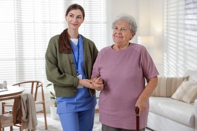 Photo of Caregiver assisting senior woman with walking cane indoors. Home health care service