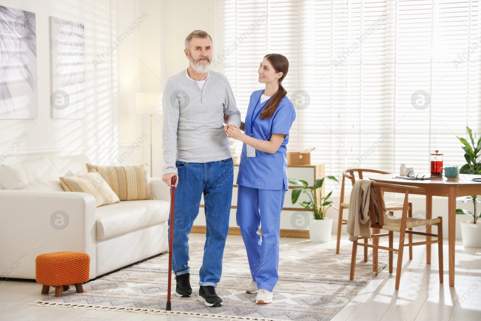 Photo of Caregiver assisting senior man with walking cane indoors. Home health care service