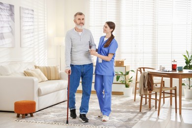 Photo of Caregiver assisting senior man with walking cane indoors. Home health care service