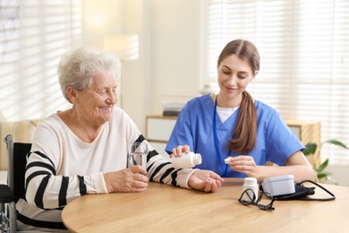 Photo of Caregiver giving pills to senior woman at table indoors. Home health care service