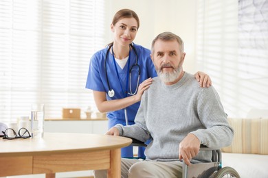 Photo of Caregiver assisting senior man in wheelchair indoors. Home health care service