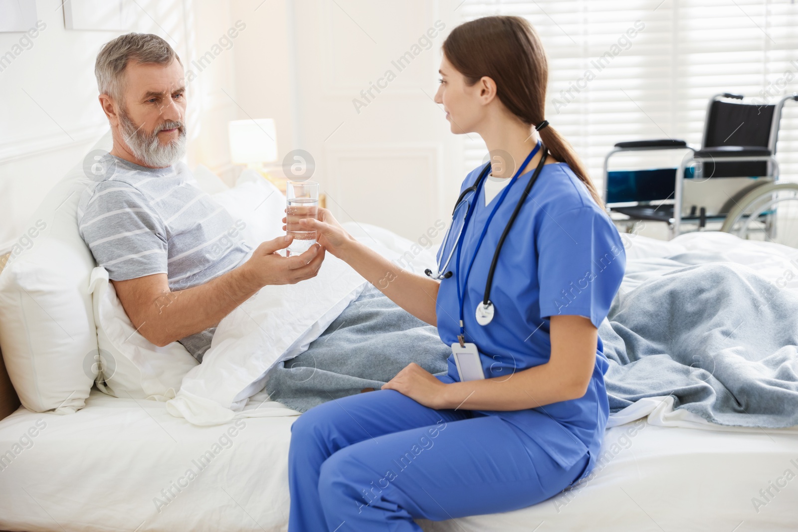 Photo of Caregiver giving water to senior man on bed indoors. Home health care service