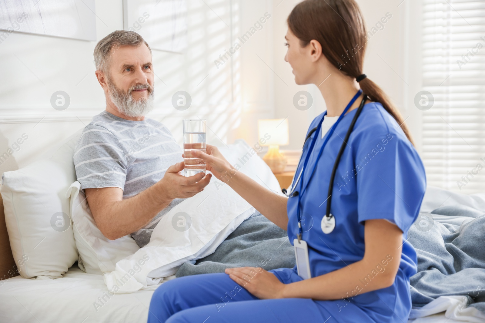 Photo of Caregiver giving water to senior man on bed indoors. Home health care service