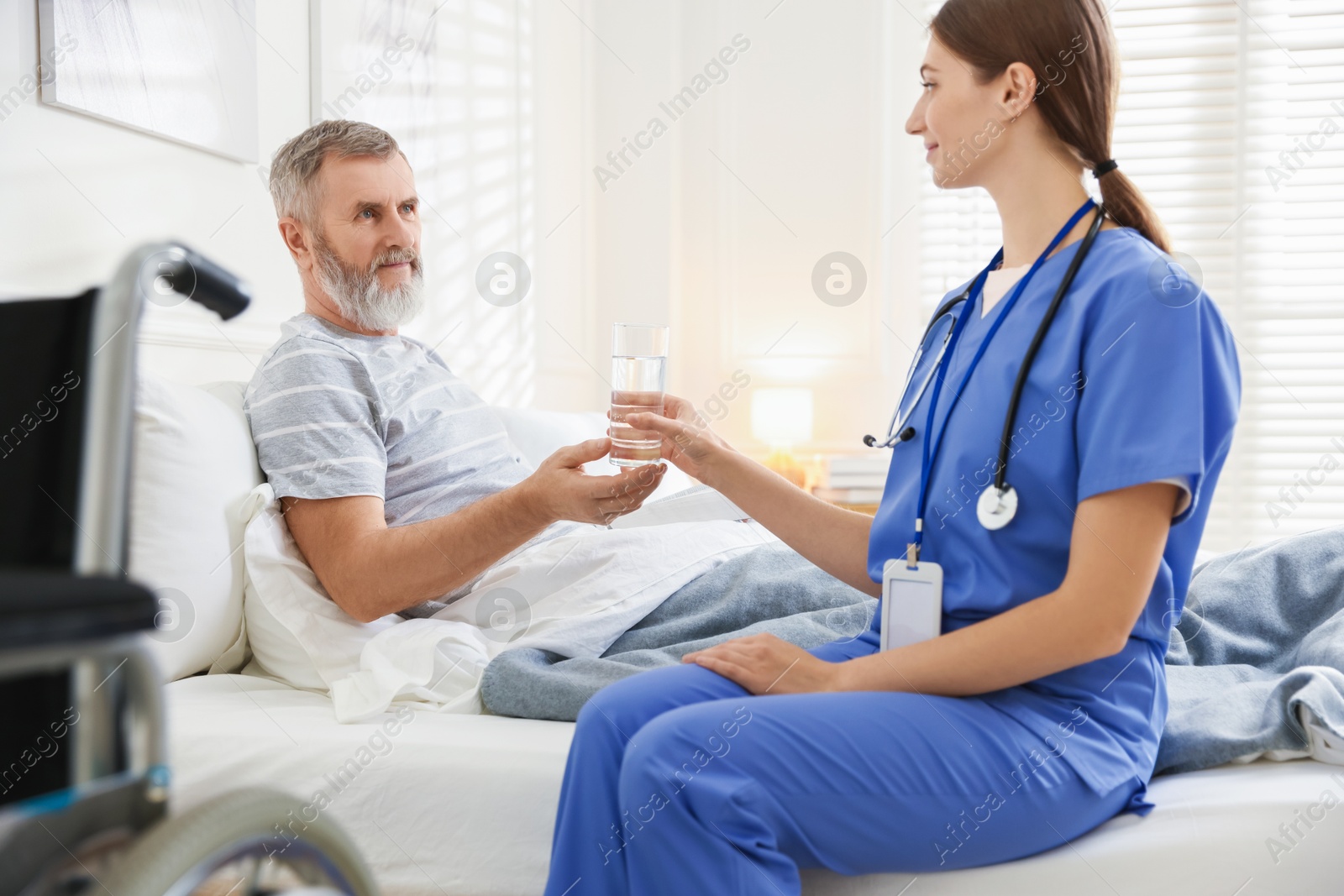 Photo of Caregiver giving water to senior man on bed indoors. Home health care service