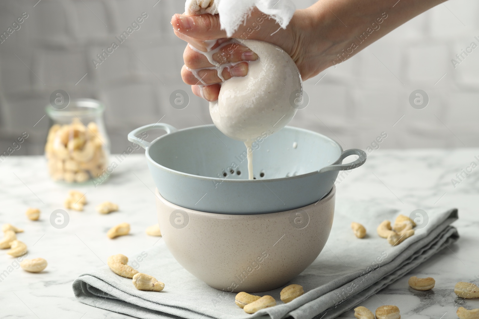 Photo of Woman straining cashew milk into colander with bowl at white marble table with nuts indoors, closeup