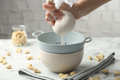 Woman straining cashew milk into colander with bowl at white marble table with nuts indoors, closeup