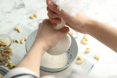 Woman straining cashew milk into colander at white table with nuts, closeup