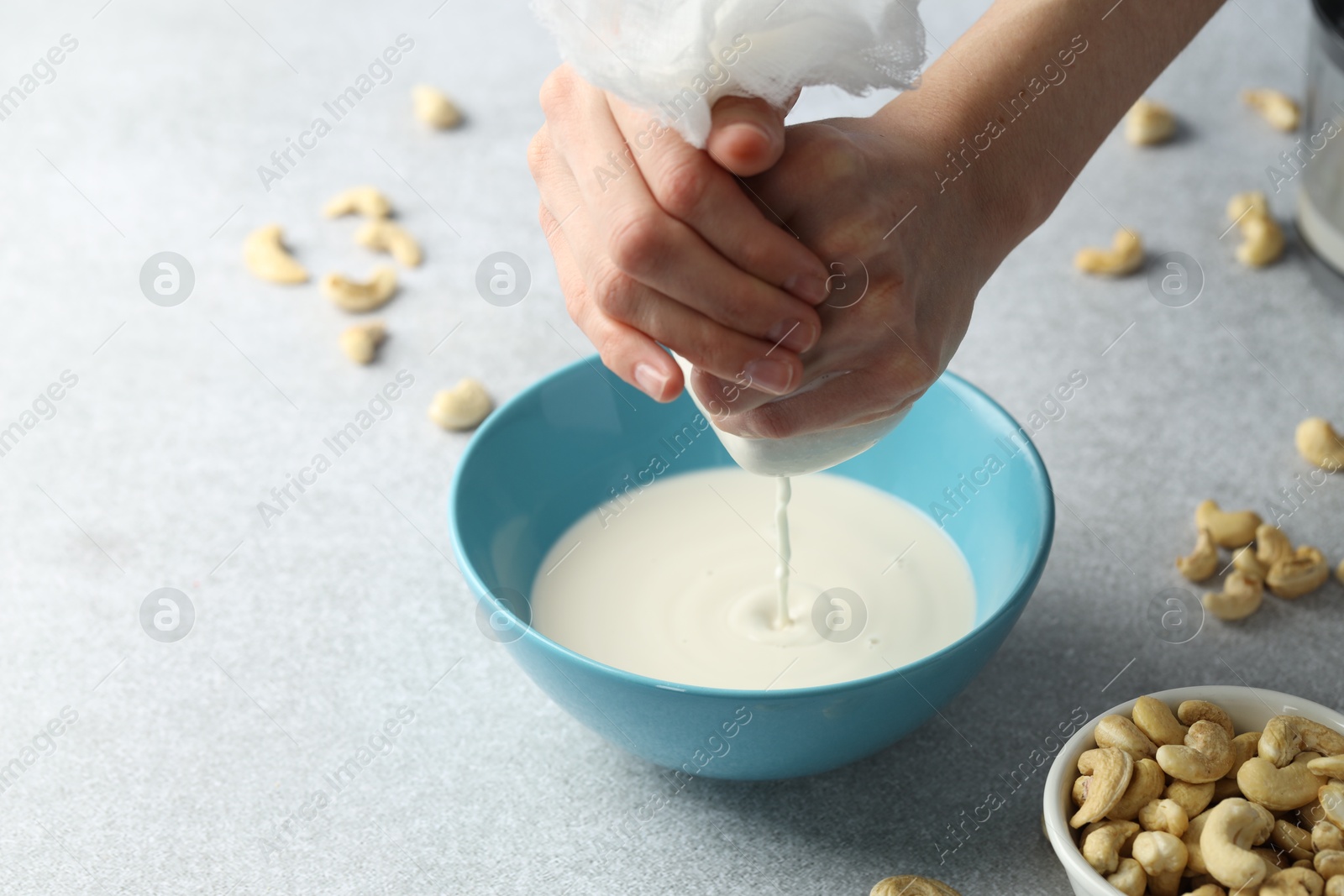 Photo of Woman straining cashew milk into bowl at light table with nuts, closeup