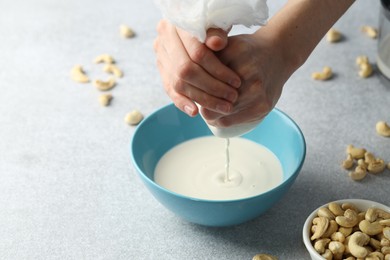 Photo of Woman straining cashew milk into bowl at light table with nuts, closeup